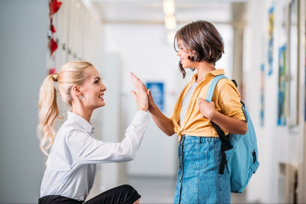 A teacher and student share a joyful greeting in a school hallway.