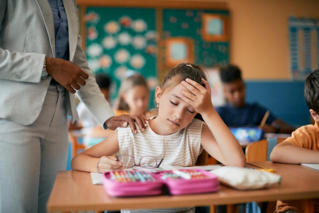 Elementary student holding her head while teacher is consoling her on a class at school. Concept of student trauma