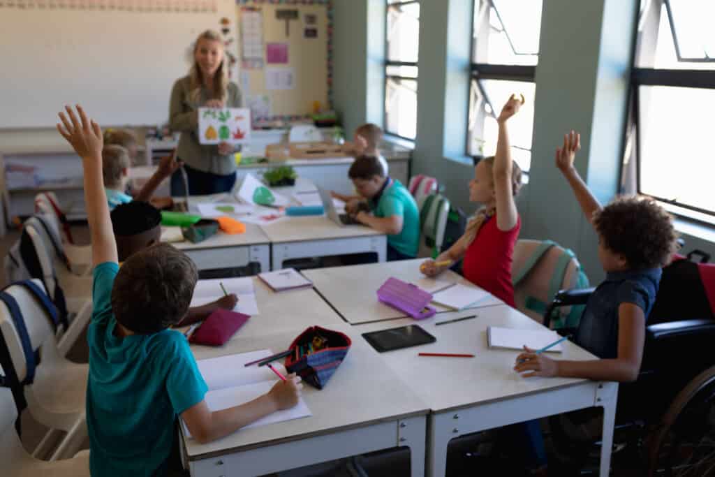 Side view of a diverse group of elementary students sitting at desks raising their hands during a lesson
