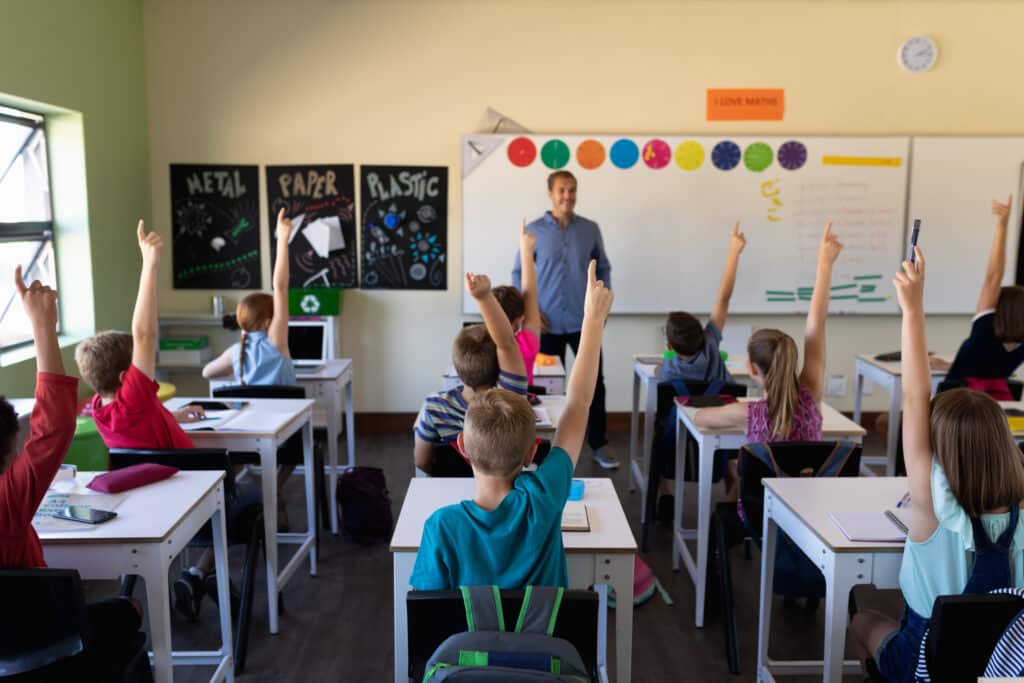 A classroom scene shows engaged students raising their hands, seated at their desks, interacting with a smiling teacher at the front. Colorful posters and a whiteboard filled with educational content about metal, paper, and plastic decorate the walls, indicating an active and enthusiastic learning environment.