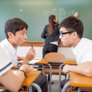 two students talking in class while teacher writes on the board